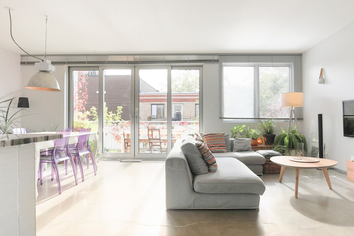 A modern living area features large glass sliding doors that offer natural light and access to the outdoor terrace. A cozy sofa is paired with a wooden coffee table, while colorful chairs adorn the dining space. Indoor plants add a touch of greenery to the environment.