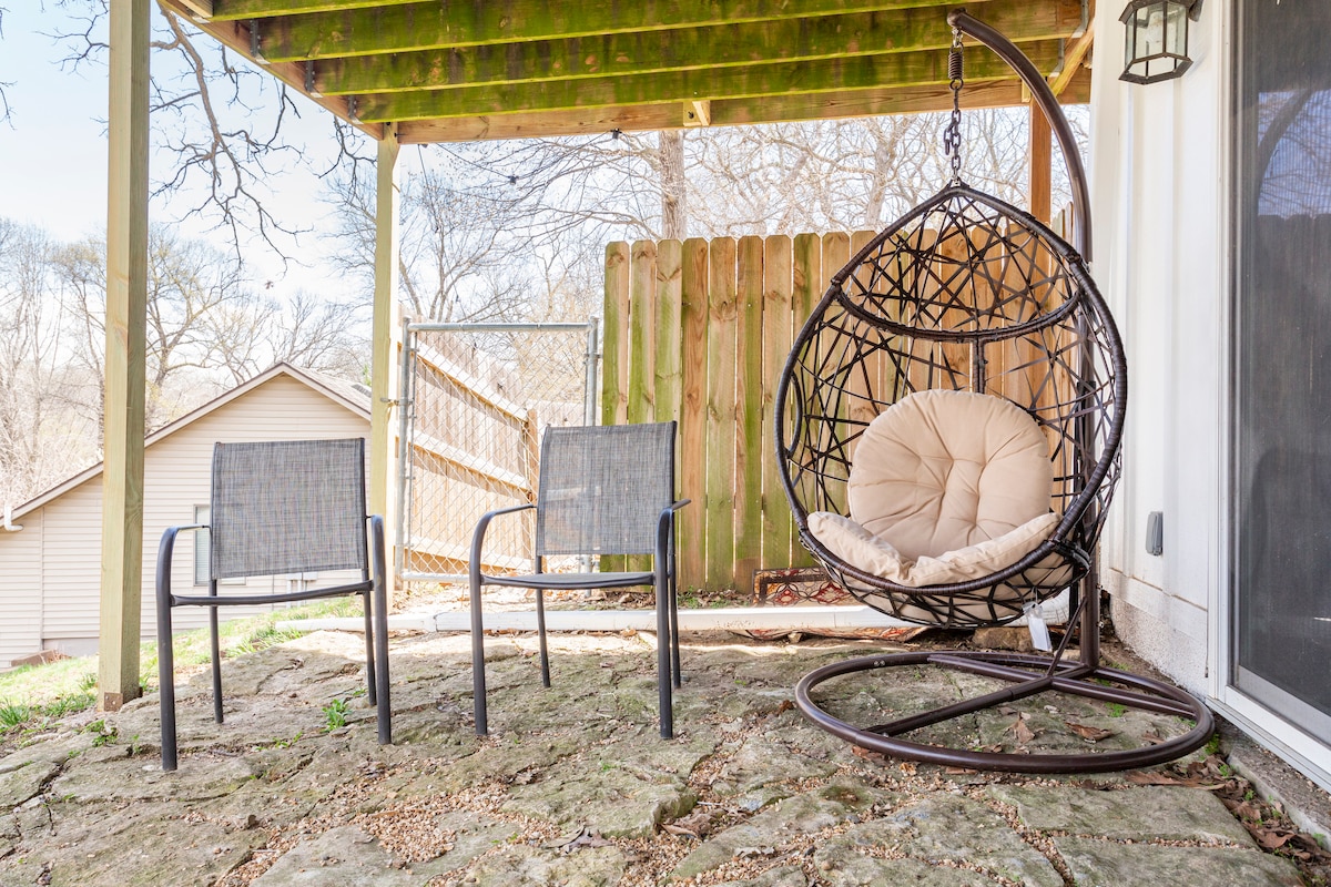 An outdoor seating area features a hanging pod chair with a cushioned seat alongside two metal chairs. The space is located under a wooden deck, with natural stone flooring and a wooden fence in the background, providing a quiet retreat.
