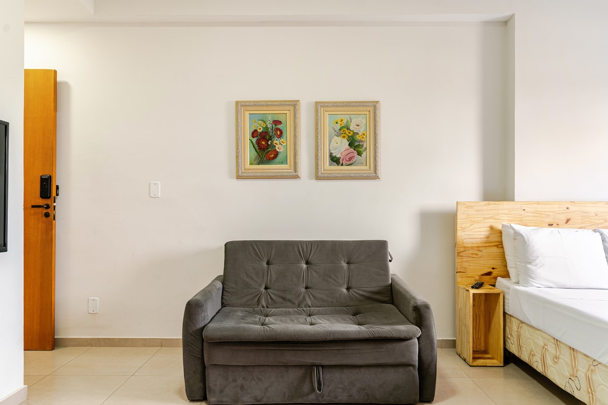 A neutral-toned space features a comfortable sofa with a pull-out option, positioned across from a simple bed. Two framed botanical artworks adorn the wall above the sofa, adding a touch of color to the room. A wooden door leads to the entrance.