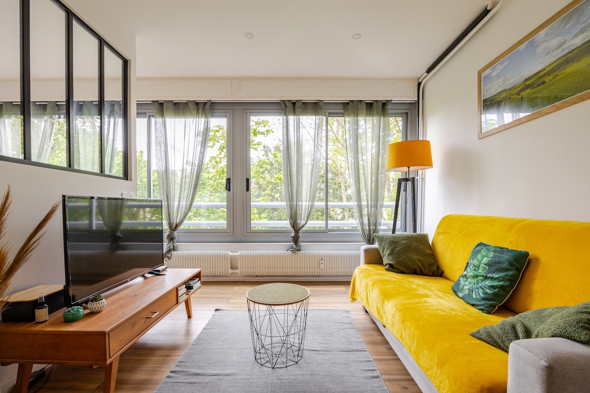 A living area features a bright yellow sofa complemented by green throw pillows. A small round coffee table is situated in front of the sofa. Large windows adorned with sheer curtains allow natural light to illuminate the space, with a scenic view of greenery visible outside.
