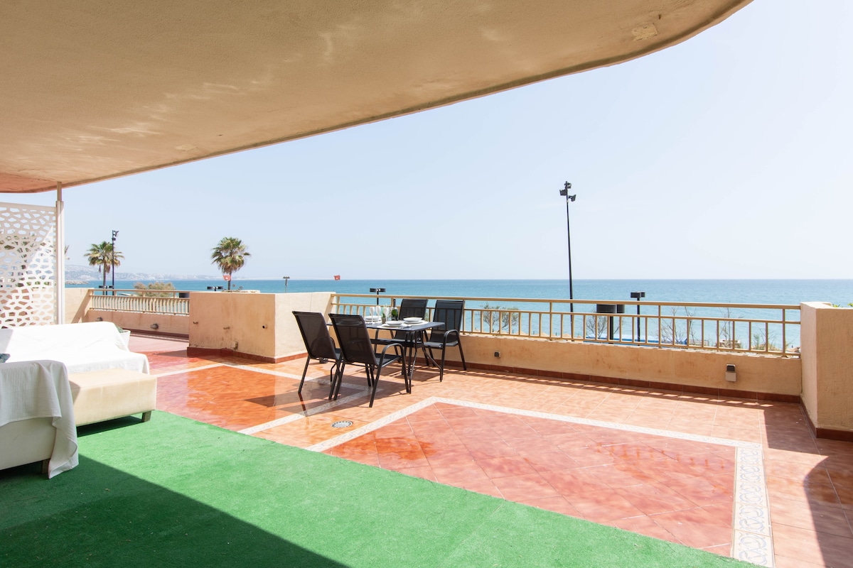 A spacious terrace offers views of the ocean, featuring a dining table with six black chairs. The area combines tile and grass surfaces, providing ample space for relaxation, while palm trees can be seen in the distance. The sun shines brightly over the serene beachfront.