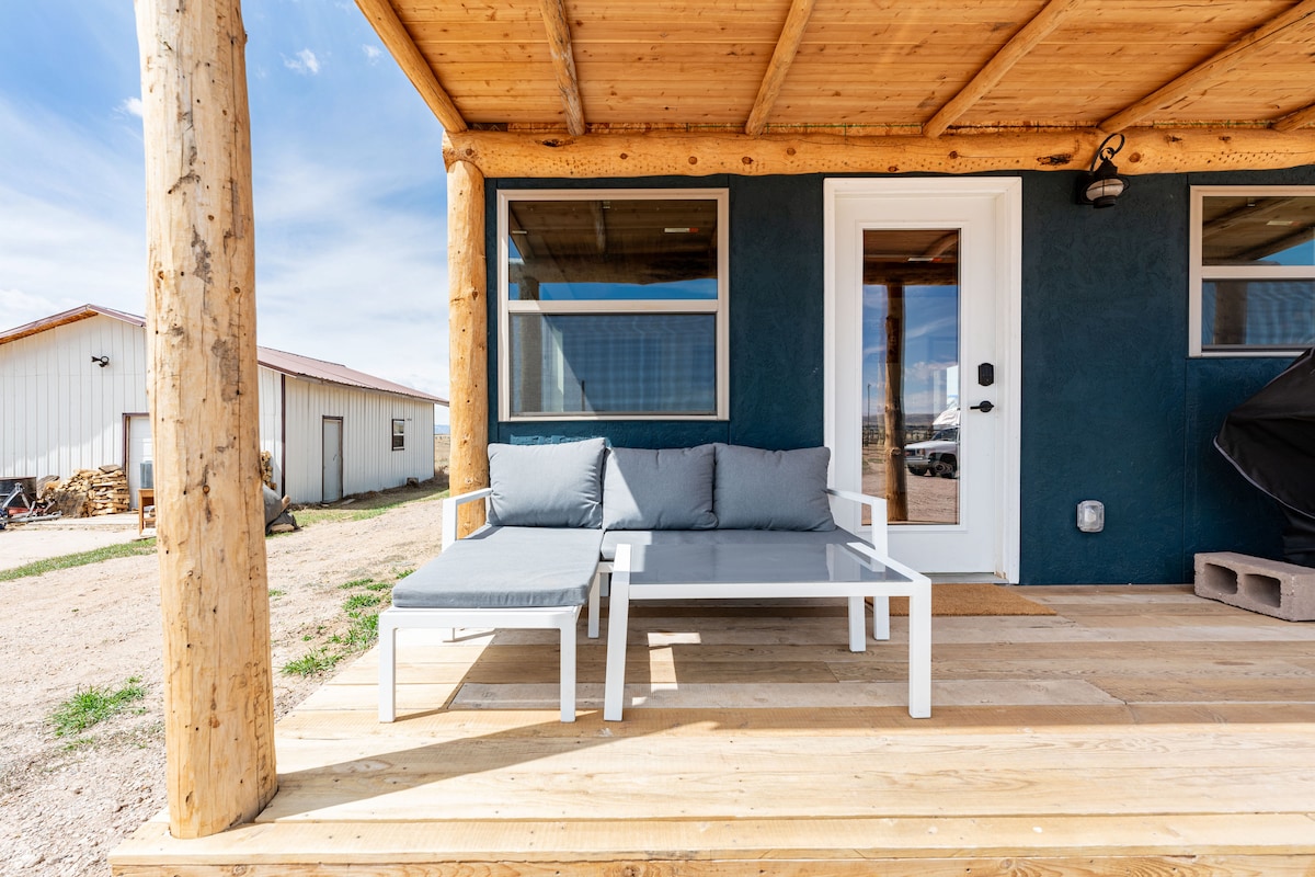 A spacious porch is highlighted with a modern white table and two gray lounge chairs, providing a comfortable outdoor seating area. Natural wooden beams support the roof, enhancing the rustic charm of the tiny home. Large windows allow views of the surrounding property.
