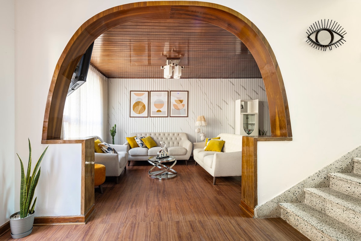 An open living area is featured, showcasing a unique archway with wooden accents. Comfortable sofas, adorned with cushions, complement a stylish coffee table. Natural light filters through sheer curtains, enhancing the warm wooden flooring and greenery present in the space.
