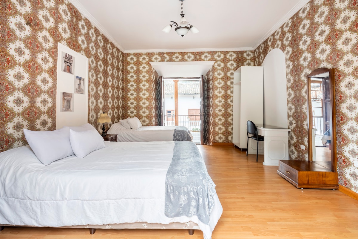 The spacious sleeping area features two beds, one made with a light coverlet and a decorative throw. A large mirror and a small desk are visible, complemented by a patterned wallpaper. Natural light enters through a sliding door that opens to a balcony.