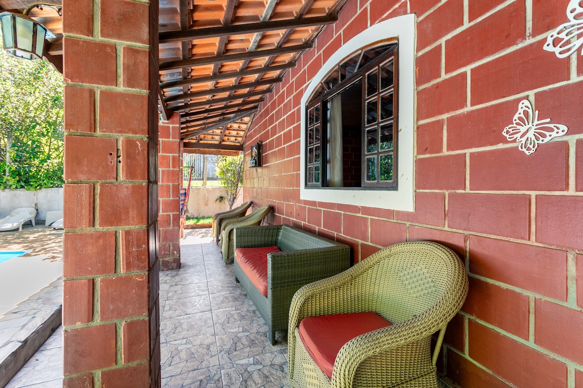 A covered patio area features two wicker chairs with red cushions, positioned near a window with wooden frames. The walls are composed of red bricks, and a tiled floor extends underneath the seating, providing a shaded space close to the pool.