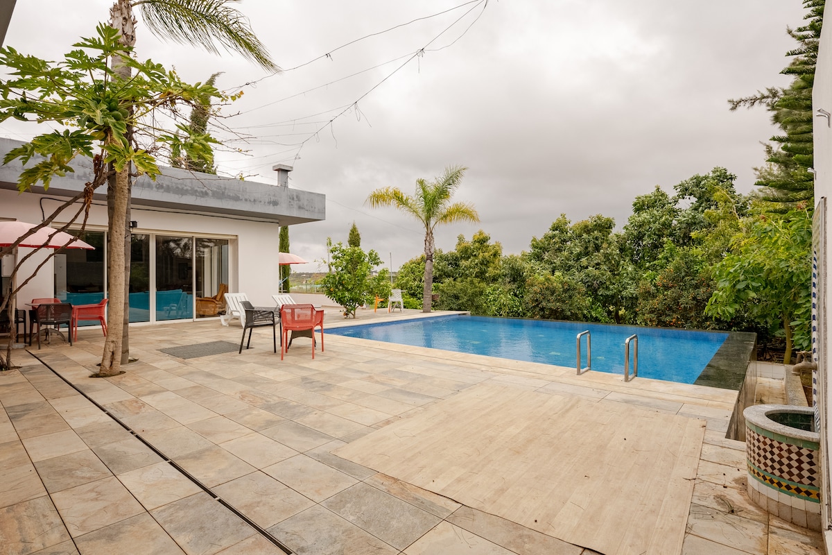 An outdoor area features a swimming pool framed by a spacious patio. Various loungers and chairs are arranged around the pool, complemented by vibrant greenery in the background. A covered seating area is also visible, providing an inviting space for relaxation.