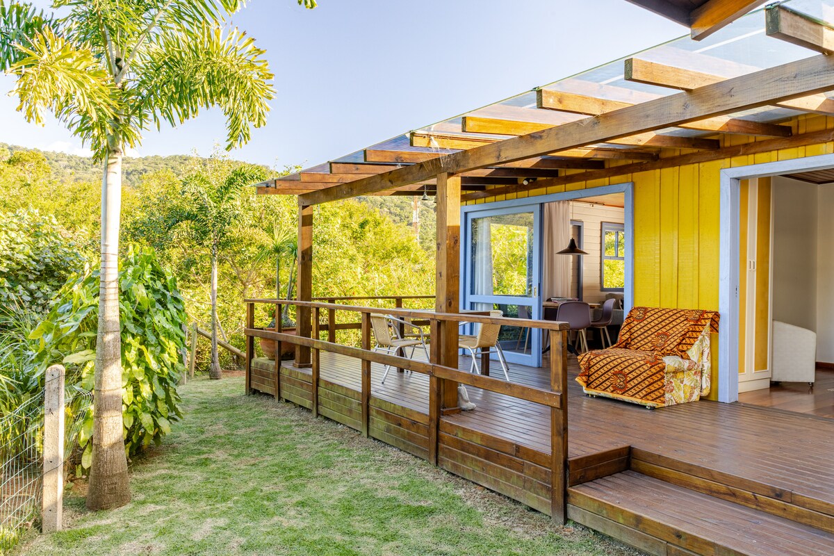A wooden deck extends from the bright yellow house, with comfortable seating arranged for relaxation. Lush greenery surrounds the area, creating a serene environment. Large sliding glass doors provide access to the interior, merging indoor and outdoor spaces harmoniously.