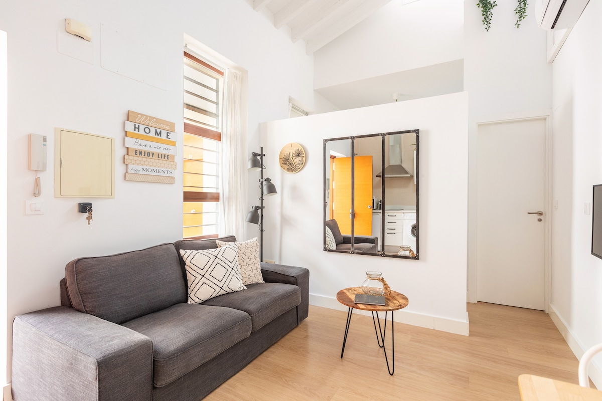 A living area features a gray sofa adorned with a patterned cushion, complemented by a small round table made of wood. A mirror on the wall reflects a glimpse of the kitchenette, while natural light fills the space, highlighting the wooden flooring and bright walls.