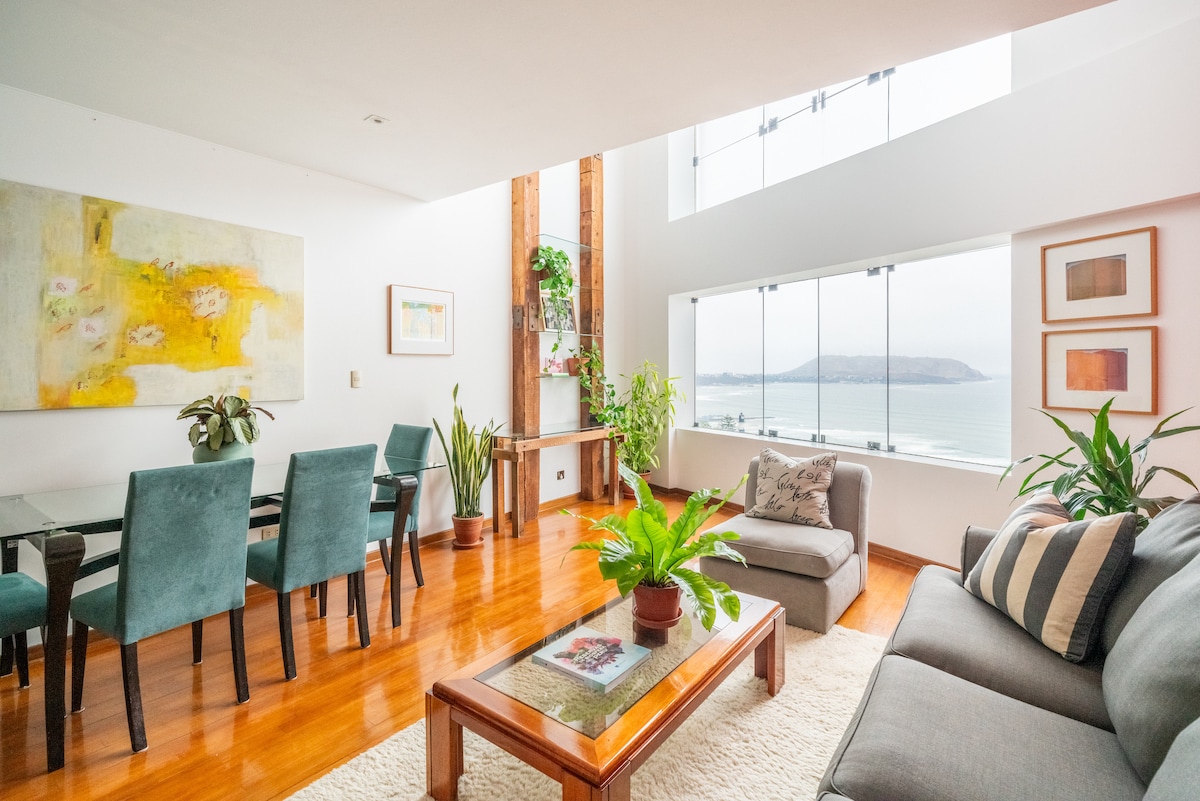 A well-lit living space features tall windows offering views of the bay. A comfortable sofa and armchair are positioned around a coffee table, while a dining area with a table and four chairs is visible. Indoor plants add a touch of greenery to the room.