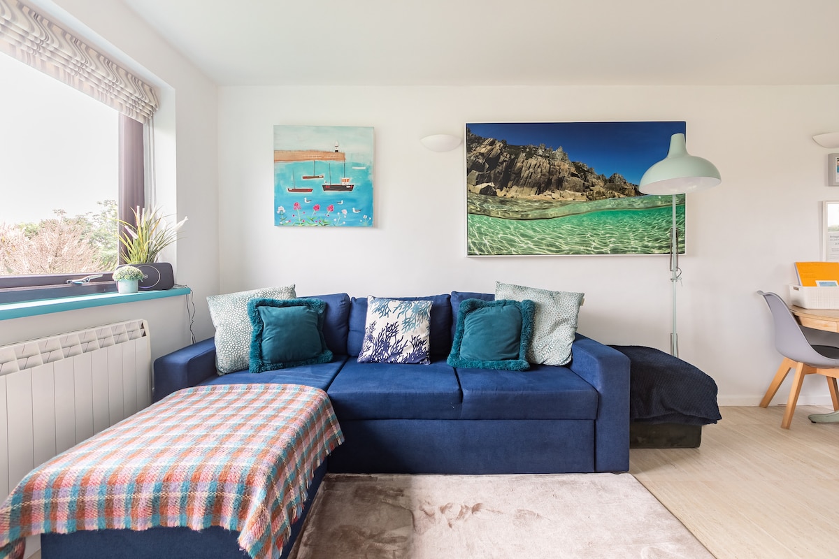 A comfortable blue sectional sofa is positioned in the living area. Colorful accent pillows enhance its appeal, with a woven throw draped casually across the side. Large windows provide natural light, while two vibrant artworks adorn the walls, depicting serene coastal scenes.