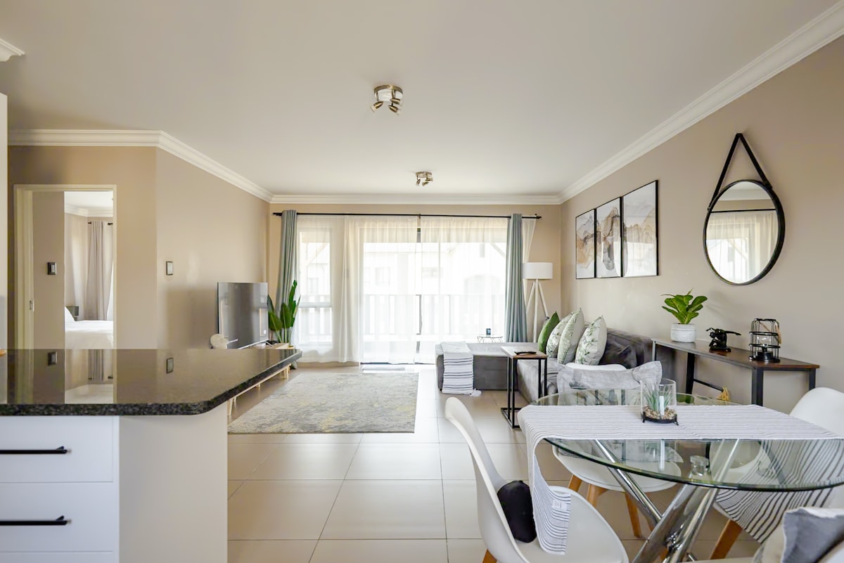 An open-concept living area is displayed, featuring a comfortable sofa and a stylish dining table with chairs. Natural light enters through large windows, which are dressed with sheer curtains. A modern television stands opposite the seating area, contributing to a welcoming environment.
