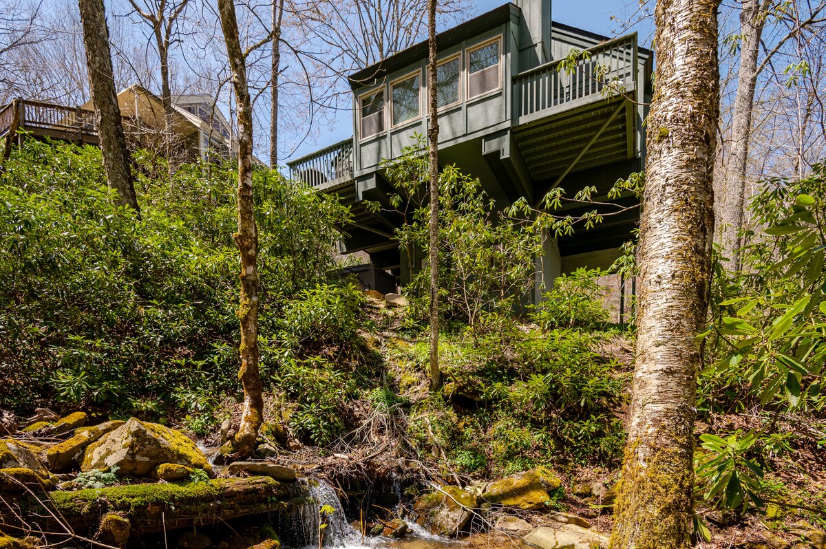 The exterior view showcases a green house elevated on pilings, surrounded by lush greenery and trees. A small stream flows nearby, adding a natural element to the landscape. The decks provide a vantage point for enjoying the peaceful outdoor setting.