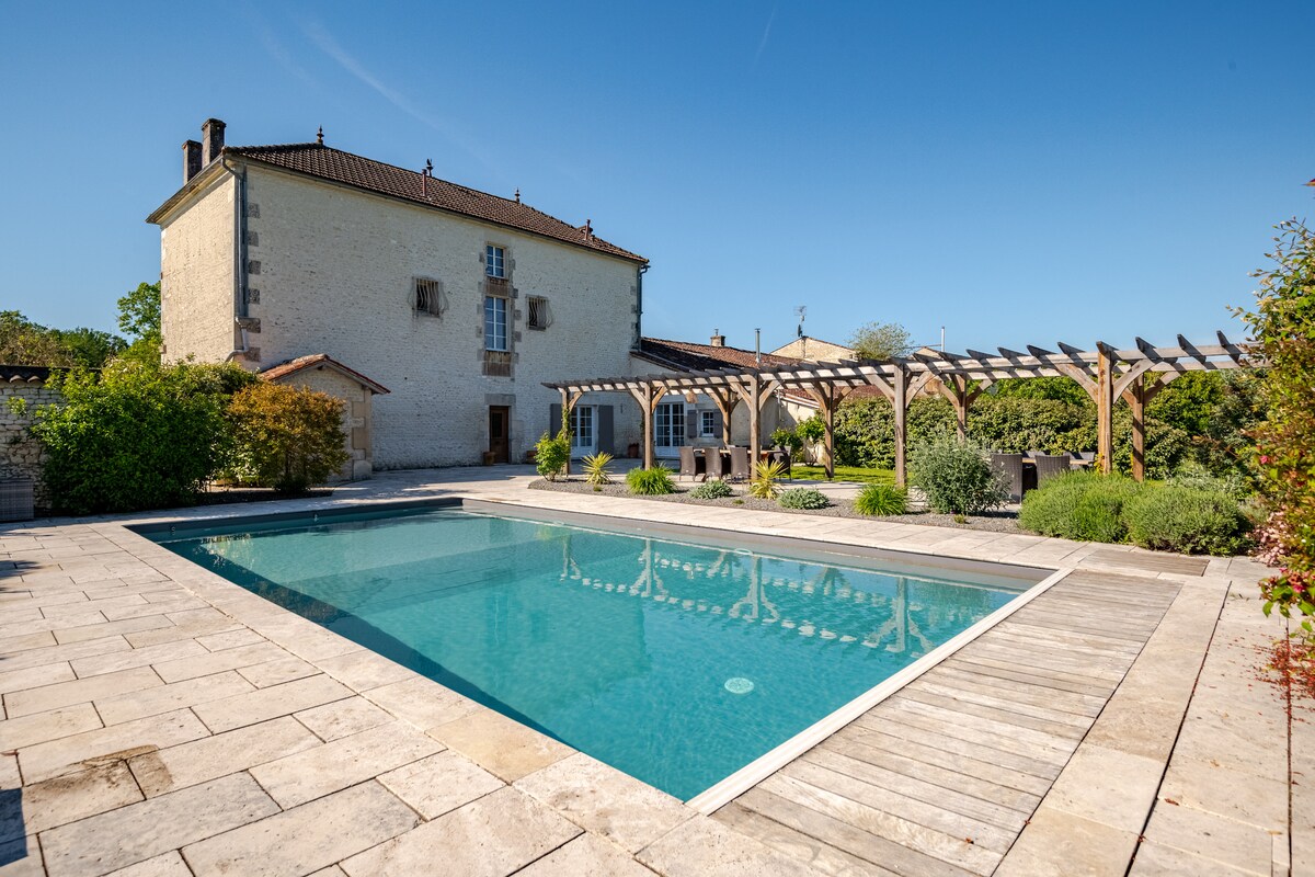 A heated swimming pool is set within a spacious terrace, surrounded by well-maintained gardens and rustic wooden pergolas. A charming two-story gite is visible in the background, showcasing traditional architecture with large windows and a light stone exterior under a clear blue sky.