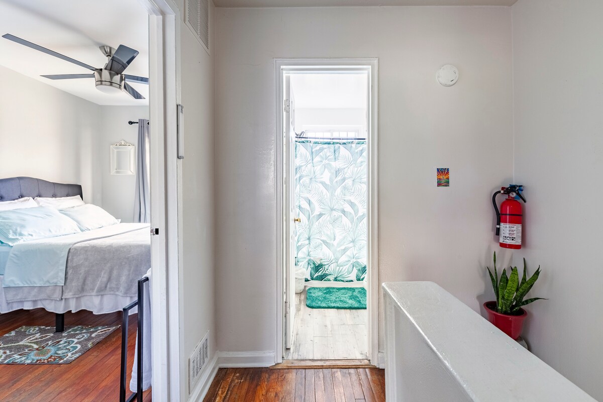 A well-lit hallway leads to a bathroom and a cozy sleeping area, with a fan overhead. Light-colored walls enhance the bright atmosphere. A fire extinguisher and a potted plant are visible, contributing to the overall safety and comfort of the space.