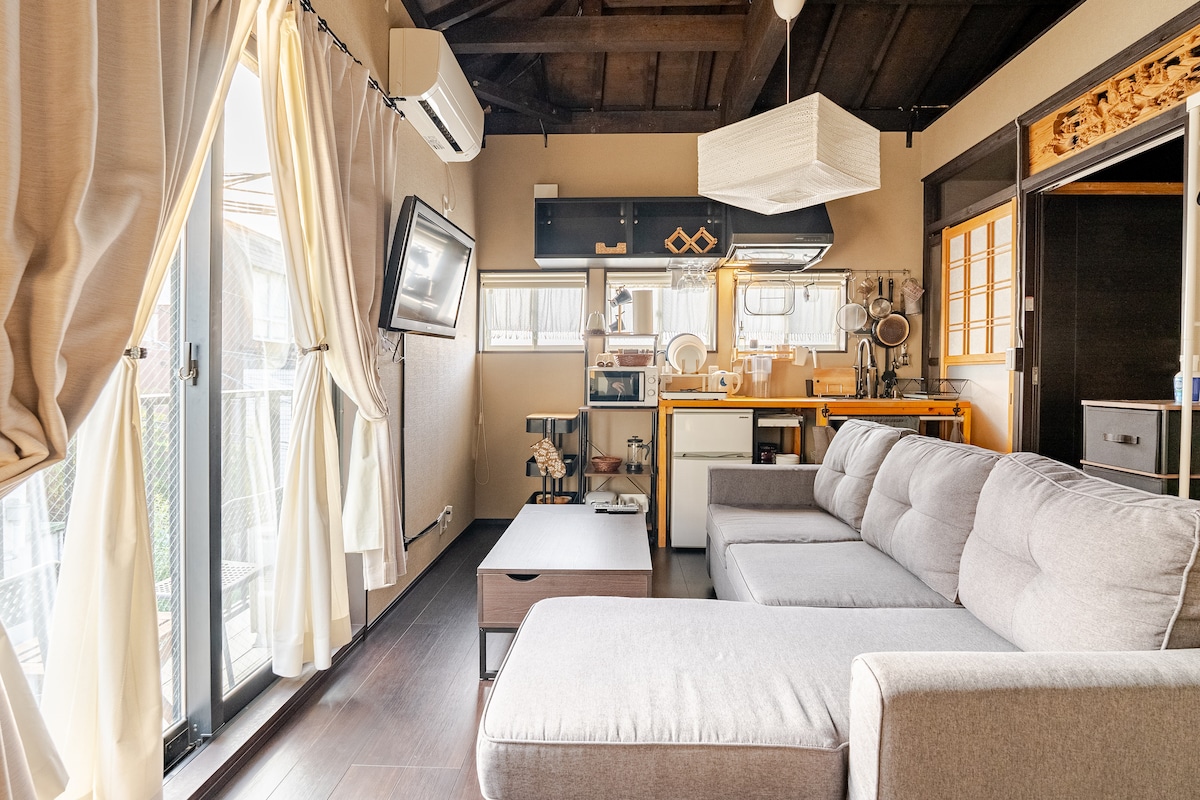 A spacious living area features an L-shaped sofa, positioned next to a coffee table. A simple kitchen, equipped with essential appliances, is visible in the background. Natural light filters through the large glass doors, highlighting a modern design accented with wood elements.
