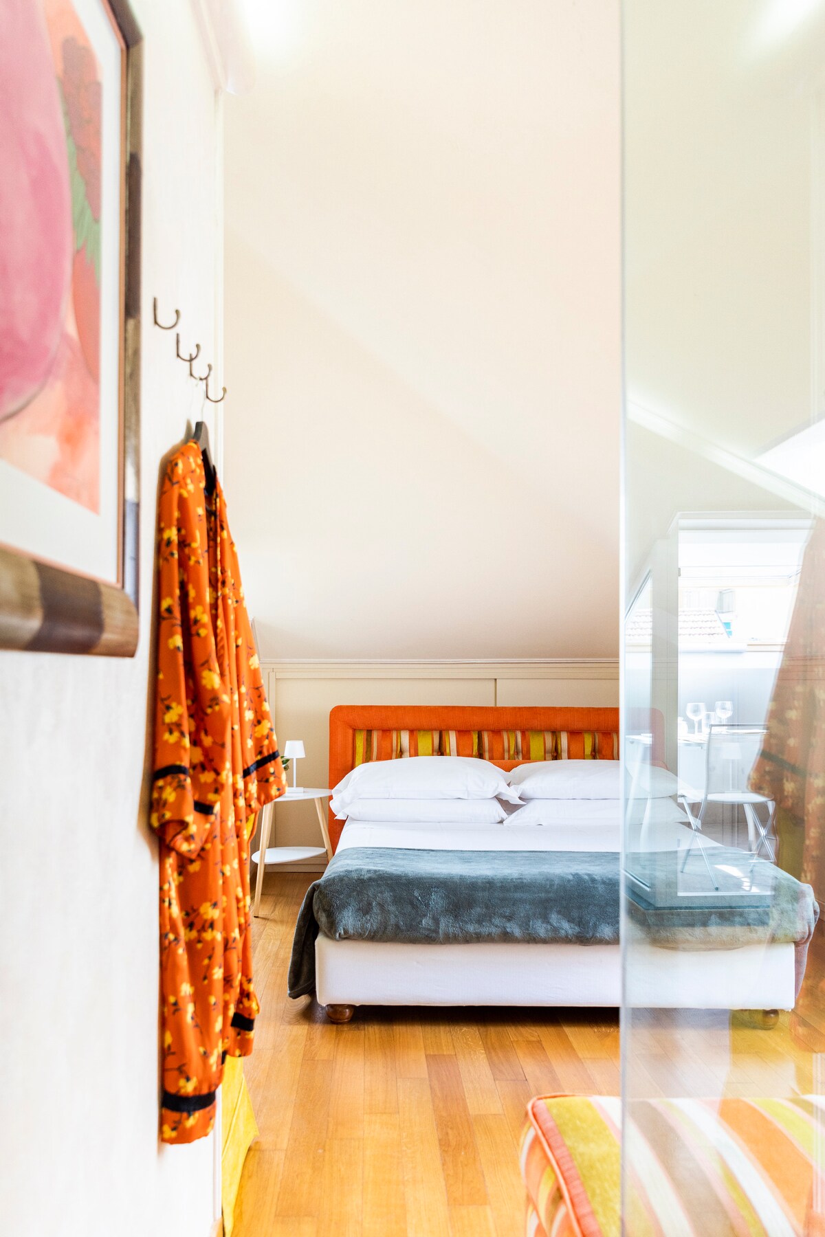 A cozy sleeping area features a spacious bed with soft linens. Colorful bedding complements the warm wooden floors. A hook displays an orange garment, adding a touch of vibrancy. Natural light floods the room through a nearby window, enhancing the inviting ambiance.