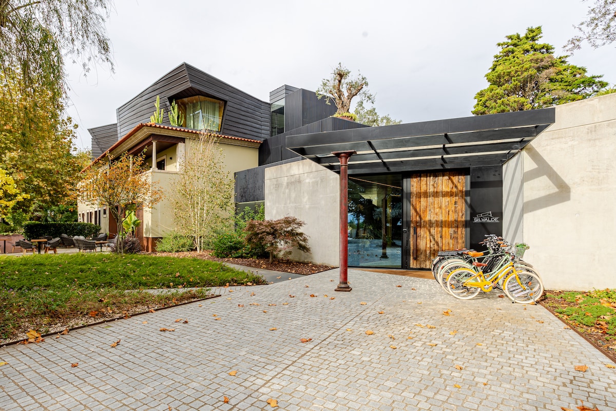 The entrance of a modern building is framed by a covered walkway and landscaped greenery. A mix of natural wood and concrete is visible, creating a harmonious look. Two bicycles are parked on the stone pathway, highlighting accessibility for guests.