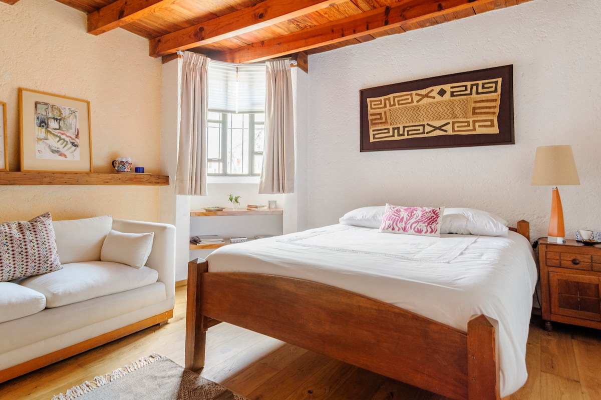 A cozy room features a spacious bed with white linens and a decorative pillow. A light-colored sofa and bedside table add functionality. Natural light enters through a window, complemented by soft drapes. Wall art and wooden beams create a warm ambiance.
