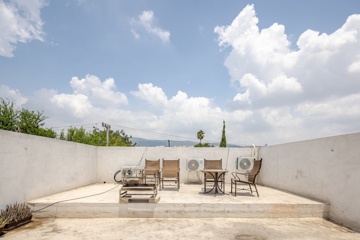 An expansive terrace is shown with a concrete surface and several lounge chairs arranged around a central table. Air conditioning units are mounted on the wall, while distant greenery is visible beyond the terrace's boundary. A clear sky with scattered clouds offers a serene backdrop.