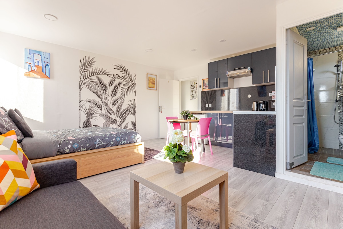 The open-plan living space features a spacious bed adorned with a patterned coverlet, and a cozy seating area with colorful cushions. A dining table with pink chairs is positioned near the modern kitchenette, which includes sleek countertops. Natural light filters through large windows, enhancing the serene atmosphere.