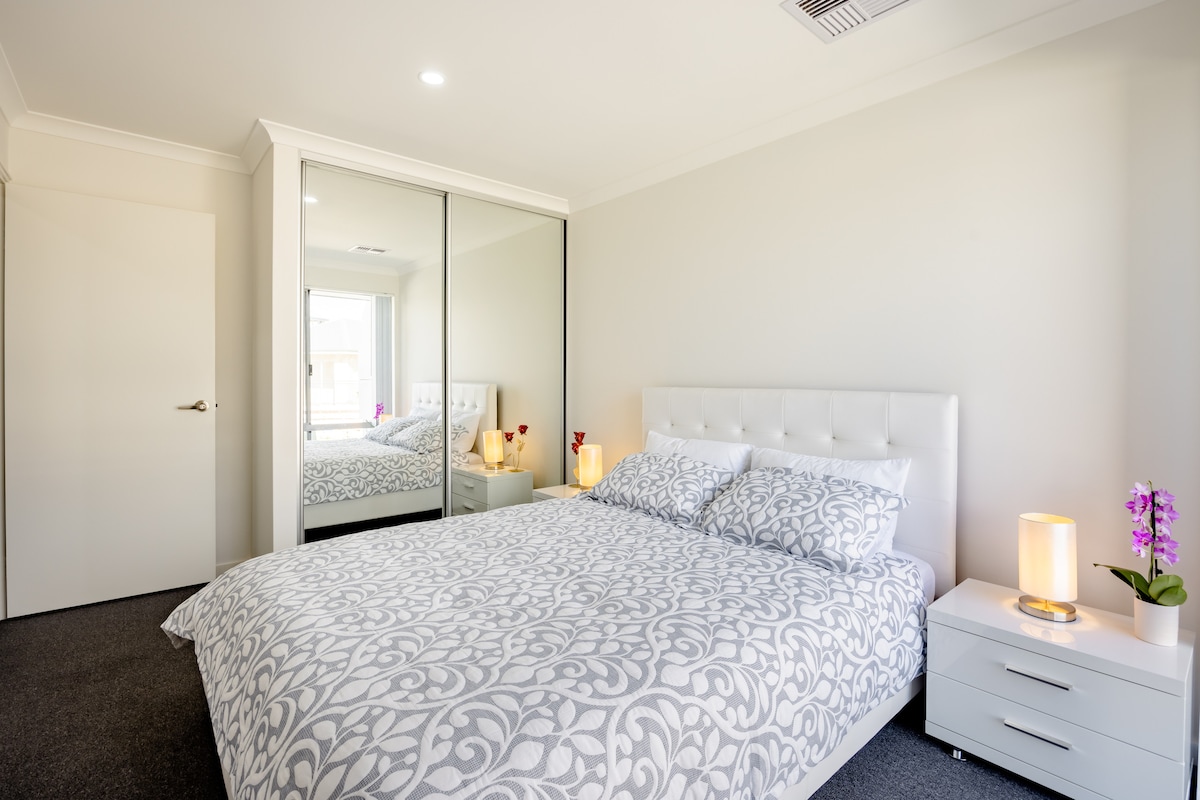 A bright bedroom features a spacious bed adorned with a patterned grey and white duvet. Mirrored wardrobes are present, reflecting the natural light from the window. A bedside table holds a lamp and a small potted orchid, contributing to the room's calming atmosphere.