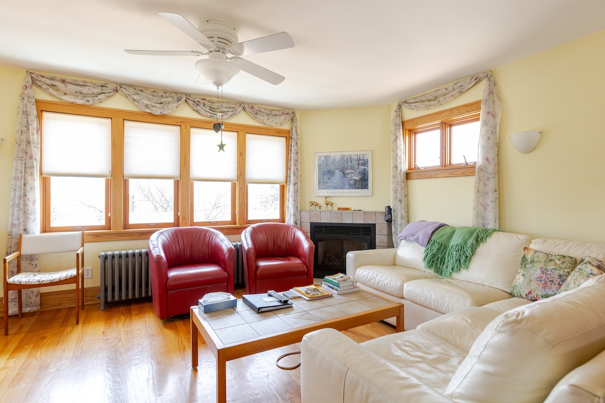 A spacious living area is filled with natural light from the multiple windows, framed by light floral drapes. Two red armchairs sit opposite a white sofa, while a wooden coffee table holds various books. A decorative piece is placed above a gas fireplace.