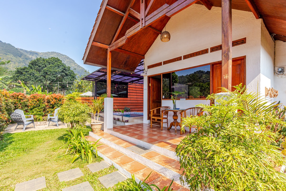 A charming outdoor seating area features comfortable chairs under a shaded porch, surrounded by lush greenery. The open space provides views of the mountains in the background, and a stone pathway leads to the entrance of the house.