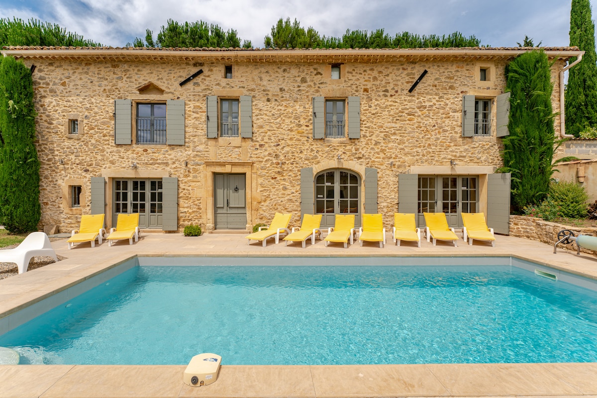 A stone house with a rustic façade is framed by lush greenery. In the foreground, a private heated pool glimmers under sunlight, surrounded by bright yellow lounge chairs for relaxation. Large windows with shutters offer views of the inviting outdoor space.