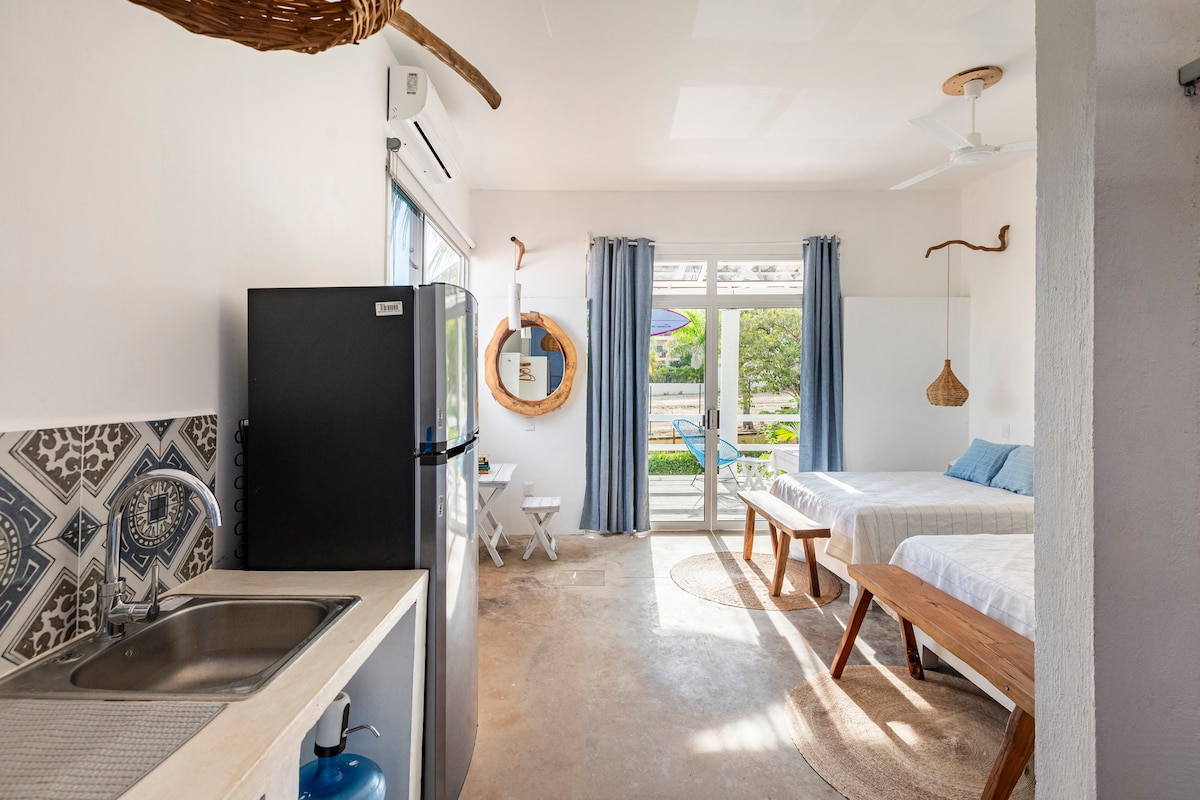The interior of the guesthouse is bright and uncluttered, featuring two comfortable double beds and a well-equipped kitchenette. The space has natural light streaming in through large glass doors, which lead to the terrace. Decorative elements include woven items and light-colored curtains.