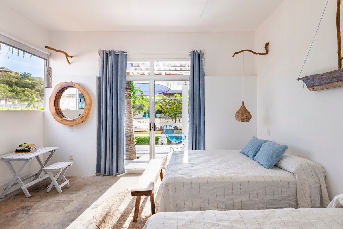 A bright and airy bedroom features two spacious beds with light linens. Large glass doors lead to a private outdoor space. Natural elements are incorporated, such as a circular mirror and driftwood decor. Soft sunlight filters in through the window, complemented by soft blue curtains.