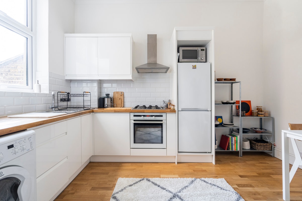A modern kitchen displays sleek white cabinetry paired with wooden countertops. A stainless steel hood and gas stove are present, alongside a microwave and refrigerator. An open shelf holds various kitchen items, while a washing machine is situated at one end. Natural light fills the space through a large window.