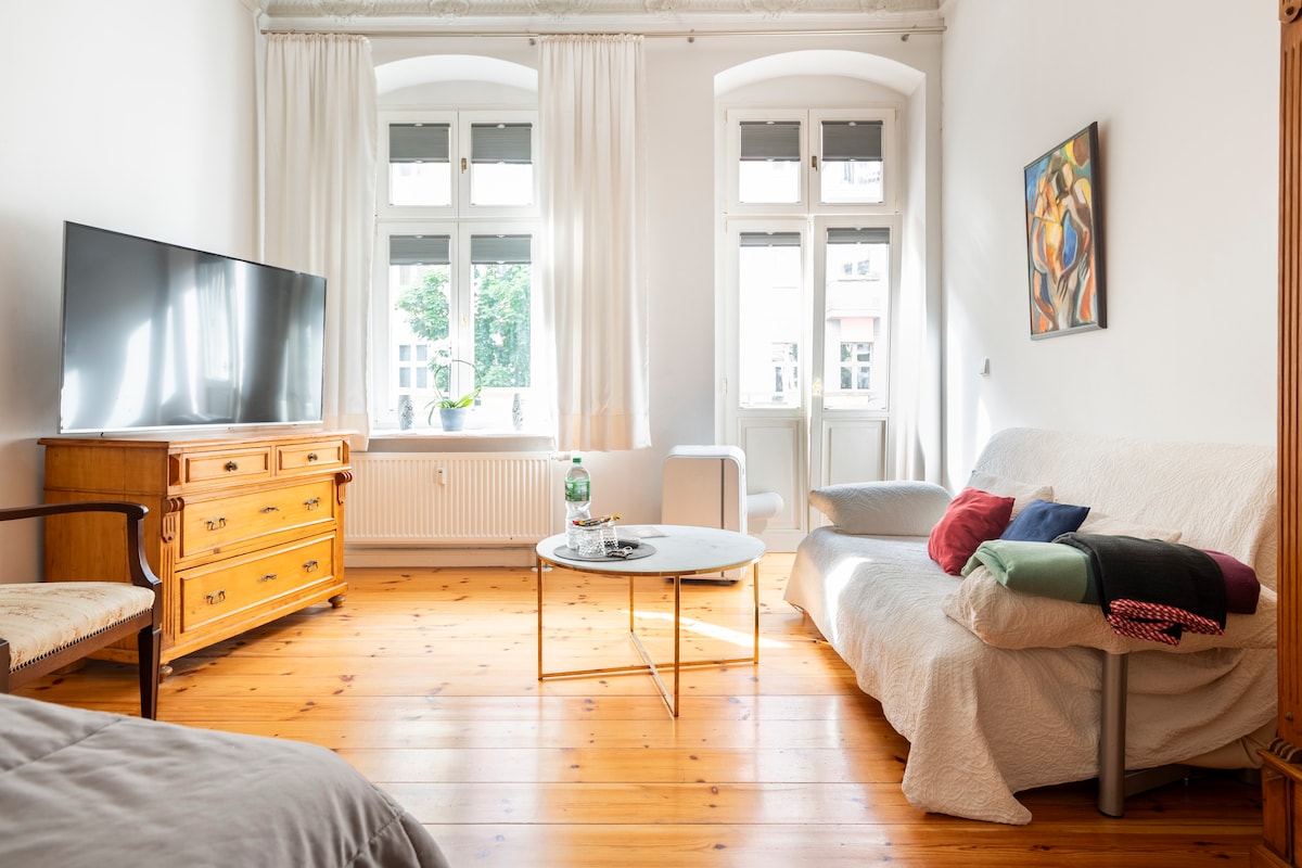 The living area features a large flat-screen television positioned against a white wall. A light wooden sideboard complements the space, alongside a coffee table with decorative items. A cozy sofa with a colorful assortment of cushions is present, and natural light streams through multiple windows covered with light curtains.