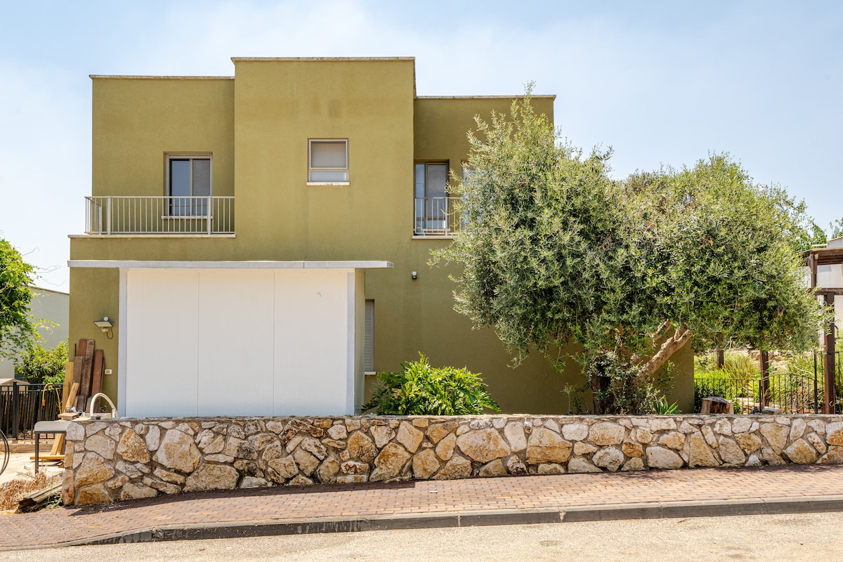 The exterior of the villa is depicted, showcasing a two-story structure with a modest green facade. A stone wall borders the property, and a shaded area with greenery can be seen in the foreground. A balcony is visible above the main entrance.