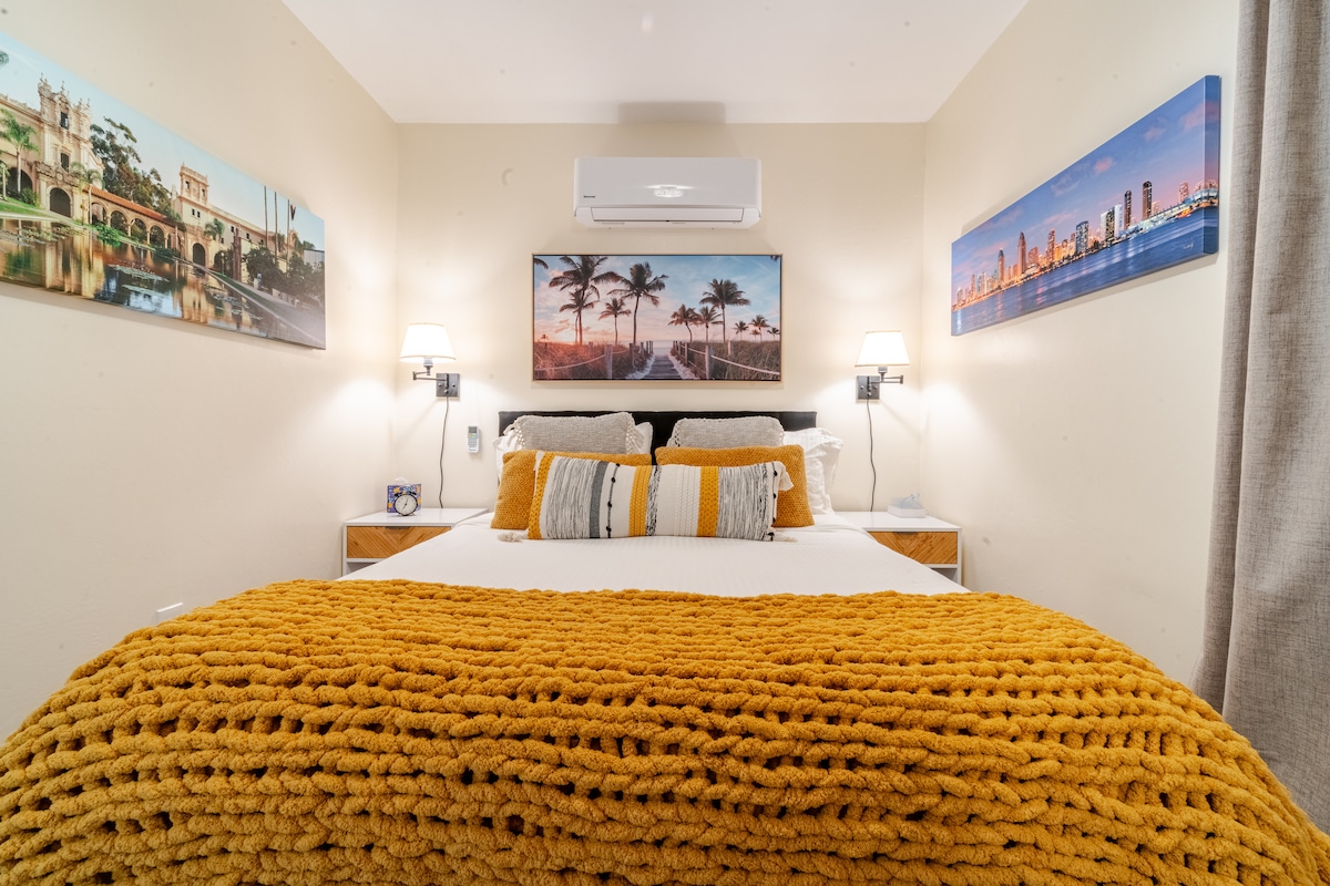 A cozy bedroom is highlighted by a spacious bed adorned with a bright yellow knit blanket. Framed photographs of palm trees and cityscapes decorate the walls, while bedside lamps provide soft illumination on either side of the bed.