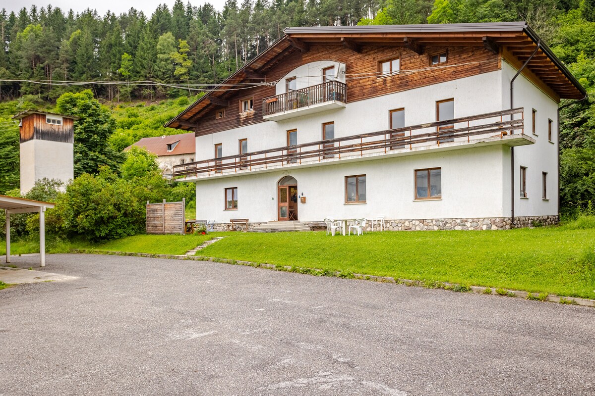 The exterior of a three-story building displays a blend of modern and traditional architectural elements. A spacious parking area is visible in the foreground, surrounded by green grass and trees. Balconies can be seen on the upper level, offering a view of the surrounding nature.