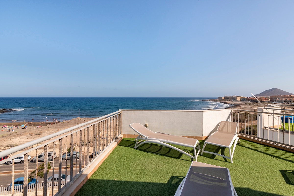 A private solarium is presented with grass flooring and two lounge chairs positioned for relaxation. The expansive view of the ocean and sandy beach is visible in the background, offering a serene backdrop for sunbathing and enjoying the outdoors.