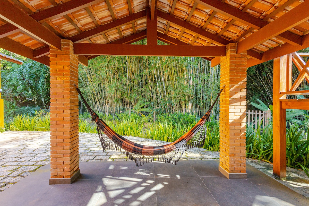 A covered outdoor space features a hammock suspended between two sturdy columns. Lush greenery and bamboo can be seen in the background, creating a serene environment. The stone floor is complemented by a patterned design, inviting relaxation in a tranquil setting.