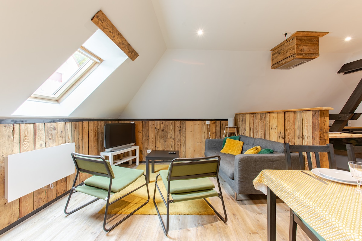 A bright living area showcases a sloped ceiling with a skylight above. Natural wood accents are present on the walls. A gray sofa and two green chairs surround a low table, complemented by a yellow rug. A television is positioned against the wooden paneling.