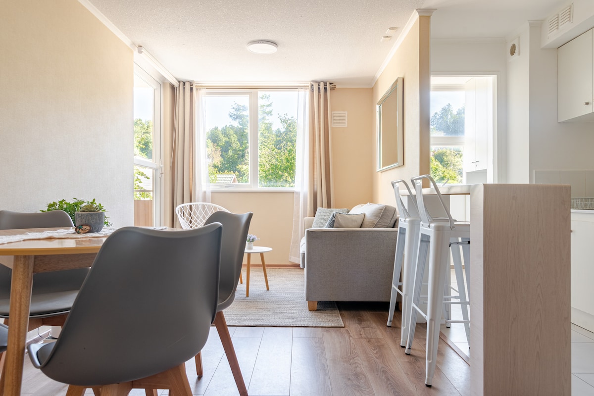 A bright living area features large windows allowing natural light to fill the space. A cozy gray sofa is paired with a dining table surrounded by four sleek chairs. Light wood flooring and soft wall colors create an inviting ambiance.