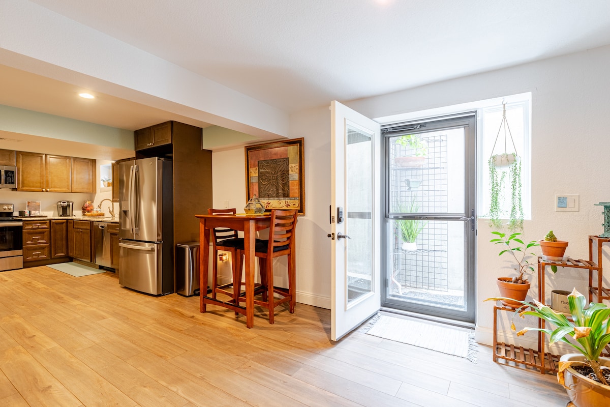 An open living area shows a kitchen equipped with modern appliances and wooden cabinetry. A dining set with four high-backed chairs is positioned near large glass doors leading to a private outdoor space. Natural light illuminates the room, enhancing its inviting atmosphere.
