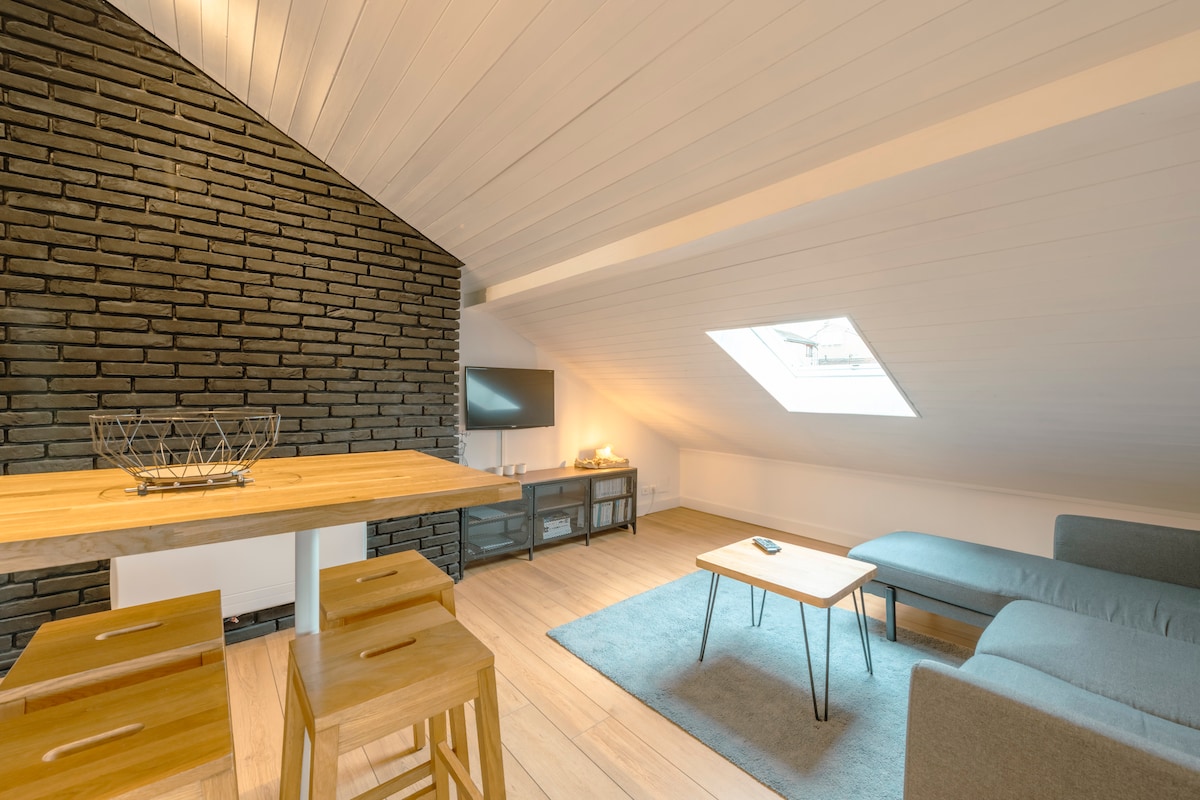 An open living space features a wooden dining table with high chairs and a low coffee table, positioned on a light wooden floor. A sleek gray couch is present, accompanied by a modern television mounted on a textured black wall. Natural light enters through a skylight.