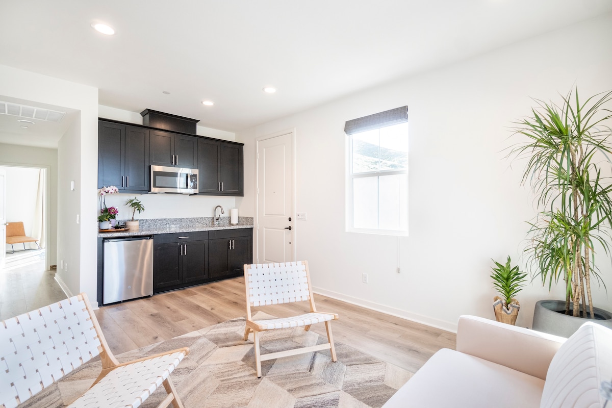 A modern living space is shown with light wood flooring and neutral walls. A sleek kitchenette, featuring a mini refrigerator and microwave, is prominently displayed. Three stylish chairs are arranged around a coffee table, while a plant adds a touch of greenery near the window.