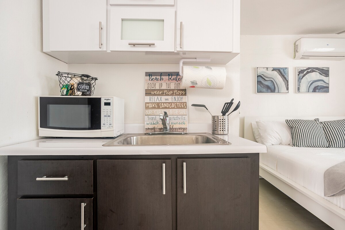 The kitchen area features modern white cabinetry with a compact sink and a microwave. A small basket and utensils are organized on the countertop. A light-colored sofa bed is visible in the background, accompanied by abstract art pieces on the wall.