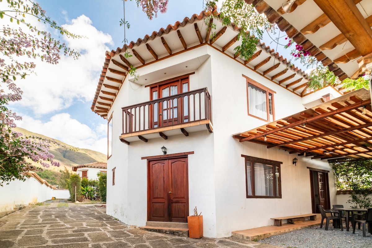 A two-story house showcases a clean, white exterior with wooden doors and a balcony. The roof is adorned with traditional tiles. Surrounding greenery adds to the serene atmosphere, while a stone pathway leads to the entrance, enhancing the inviting appearance of the home.