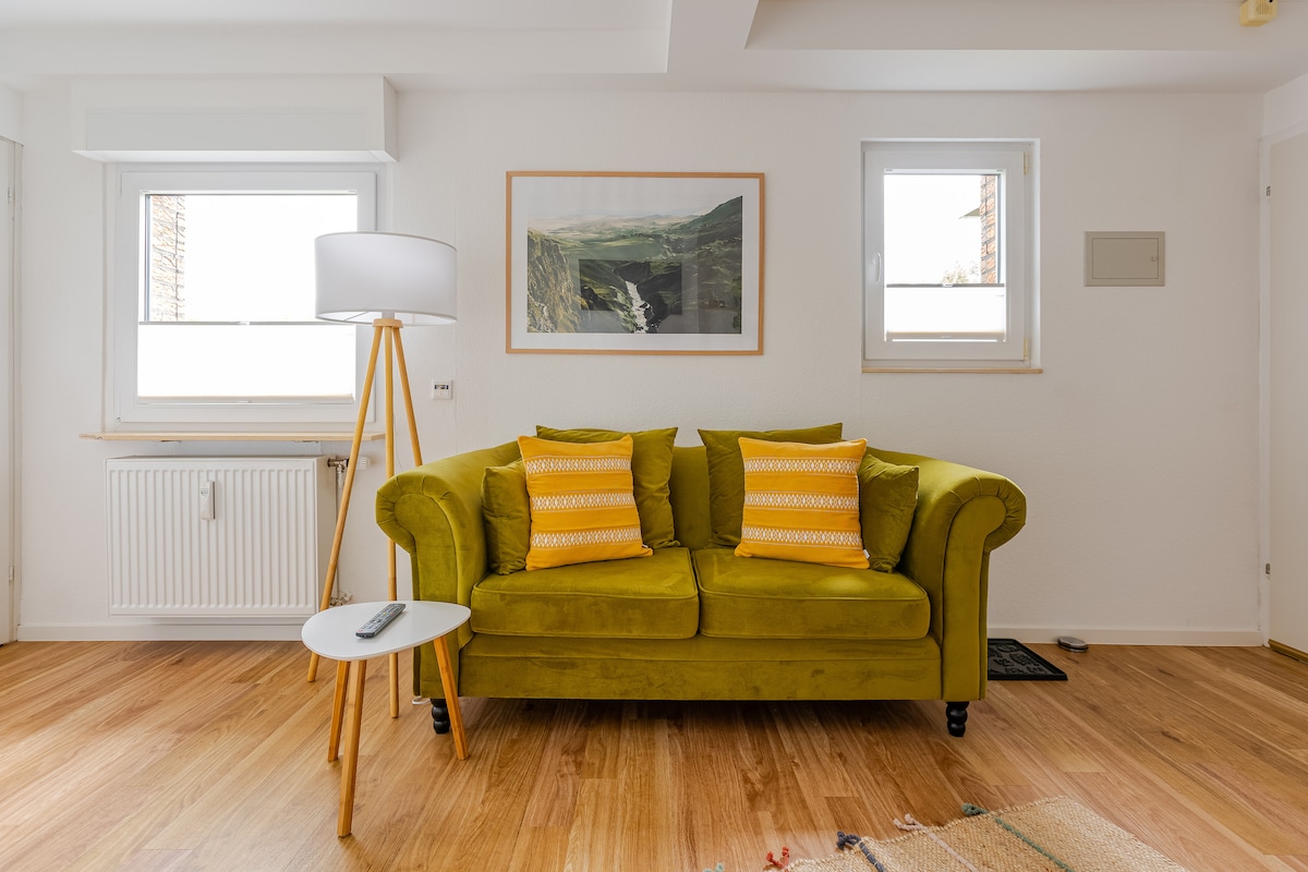A bright seating area is furnished with a green couch adorned with yellow patterned cushions. A small side table is positioned beside it, and two windows allow natural light to fill the space, complementing the light wood flooring.