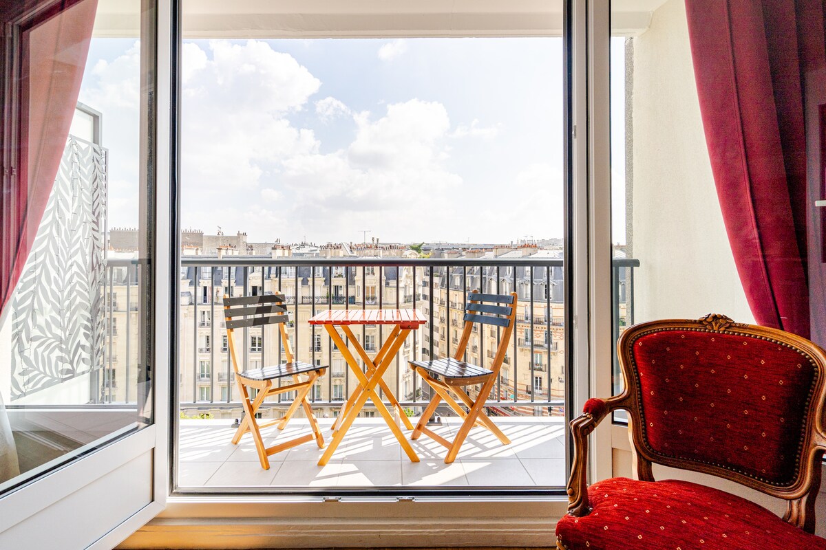 A balcony view is captured through large glass doors, featuring a small wooden table with two folding chairs. The railing showcases a cityscape in the distance, while red curtains frame the scene, inviting natural light into the interior space.