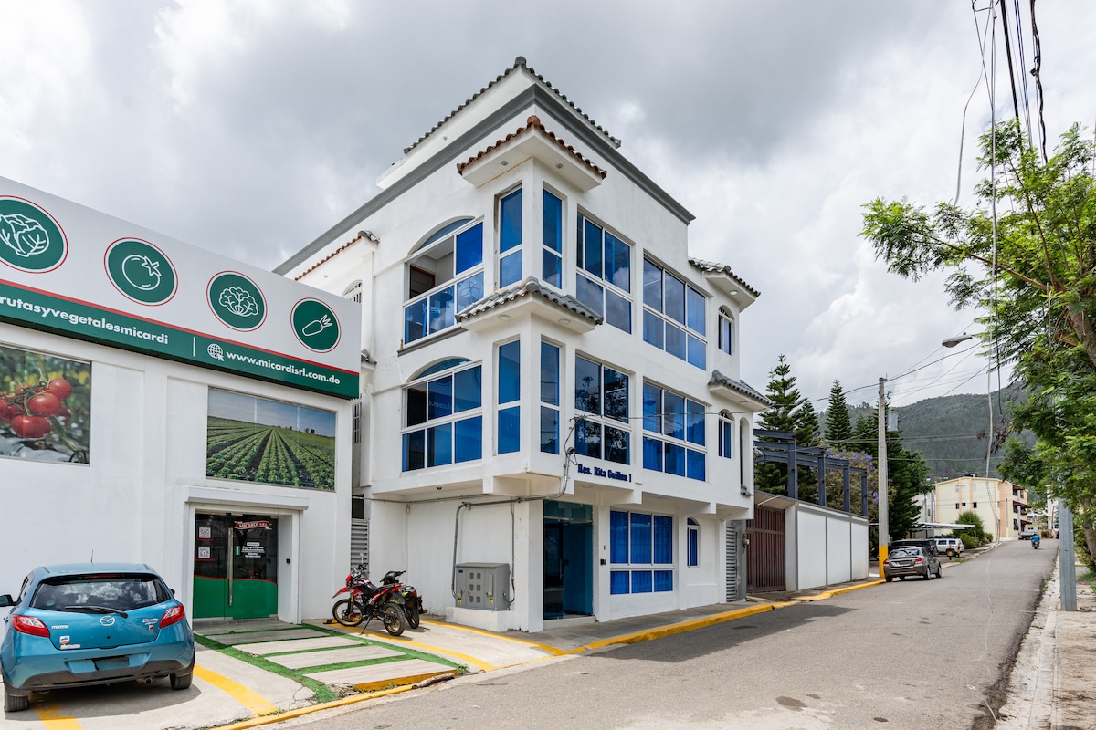 A multi-story building features large blue-framed windows that allow ample natural light. The ground floor houses a storefront with signs displaying fruits and vegetables. A vibrant blue vehicle and a motorcycle are parked outside, along a tree-lined street.