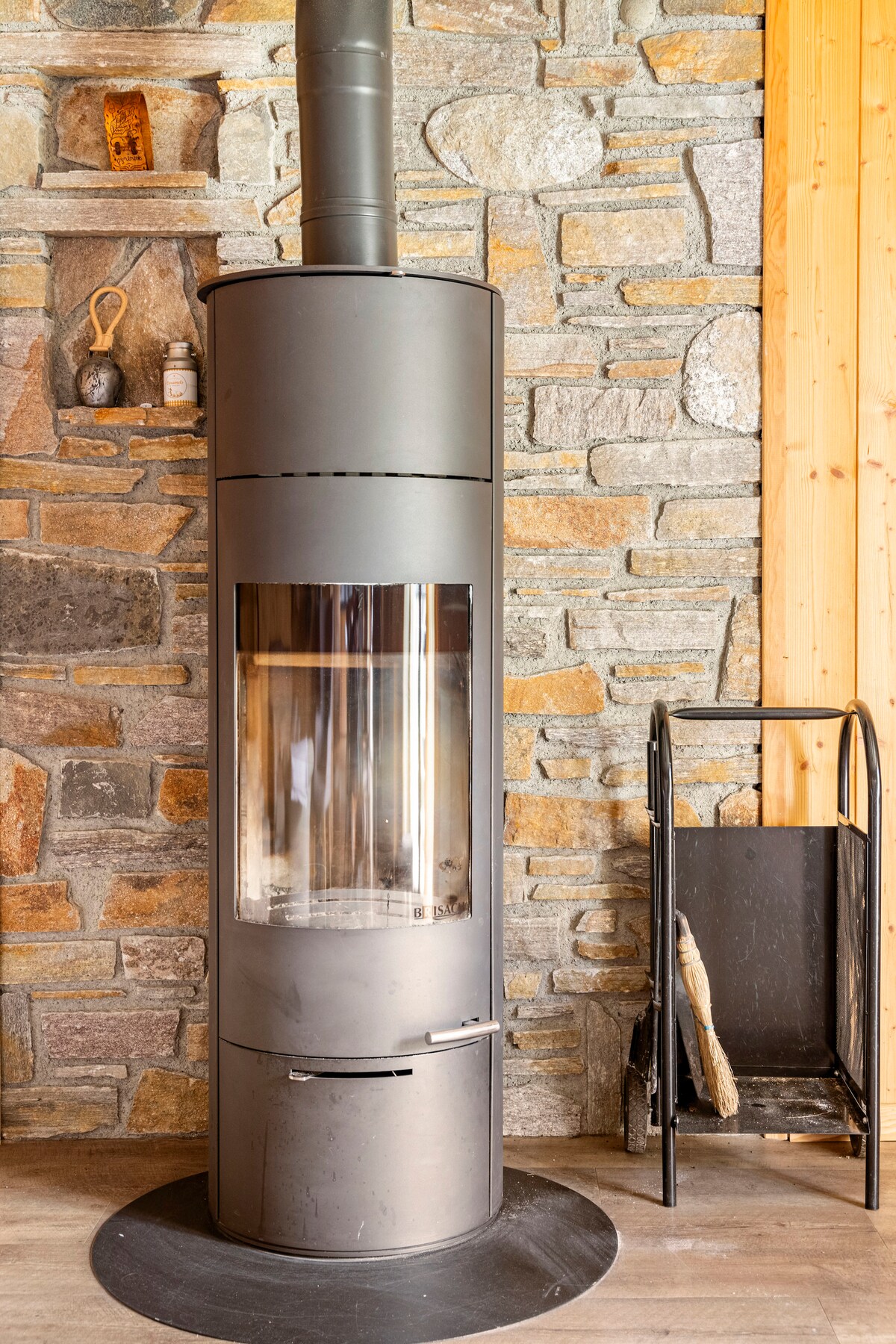 A modern wood stove is positioned against a stone wall, providing warmth and a focal point. The stove is cylindrical with a glass front, revealing a view of the flames. Next to it, a simple metal log holder stands ready for use.