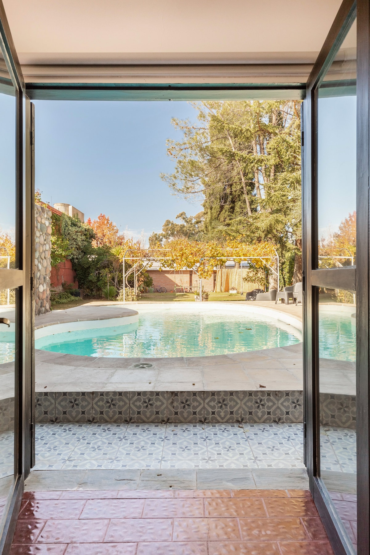 A view of a secluded swimming pool is framed by expansive glass doors. The pool features a curved design and is surrounded by trees and autumn foliage. Light reflects off the water, creating a serene atmosphere for relaxation.