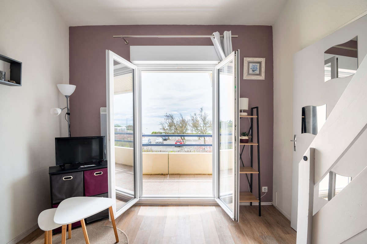A cozy living area is displayed with large glass doors opening to a balcony. A small television is positioned on a stand beside a two-drawer unit. A bright lamp and a slender bookshelf add functionality to the space, while light-colored walls complement the warm wood flooring.